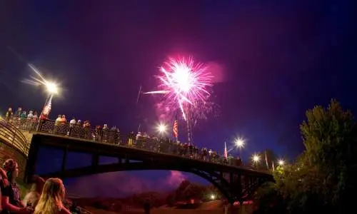 Persone sedute in un parco accanto a un ponte, che guardano i fuochi d'artificio nel cielo