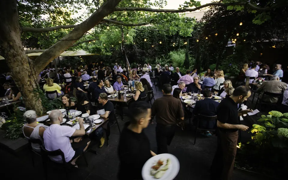 Un'affollata area pranzo all'aperto al tramonto, con tavoli tra gli alberi