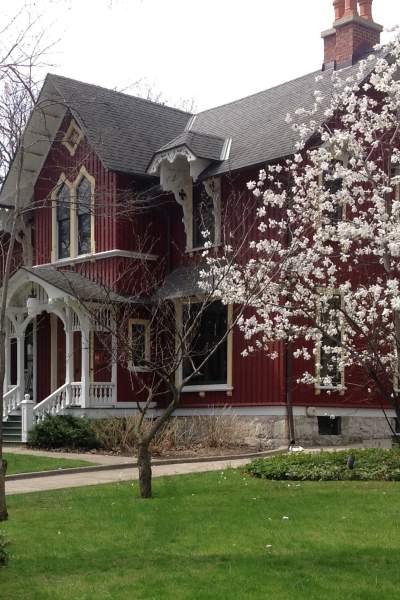 Un'imponente casa in legno rosso del XIX secolo, a due piani, dietro un prato e un albero in fiore.