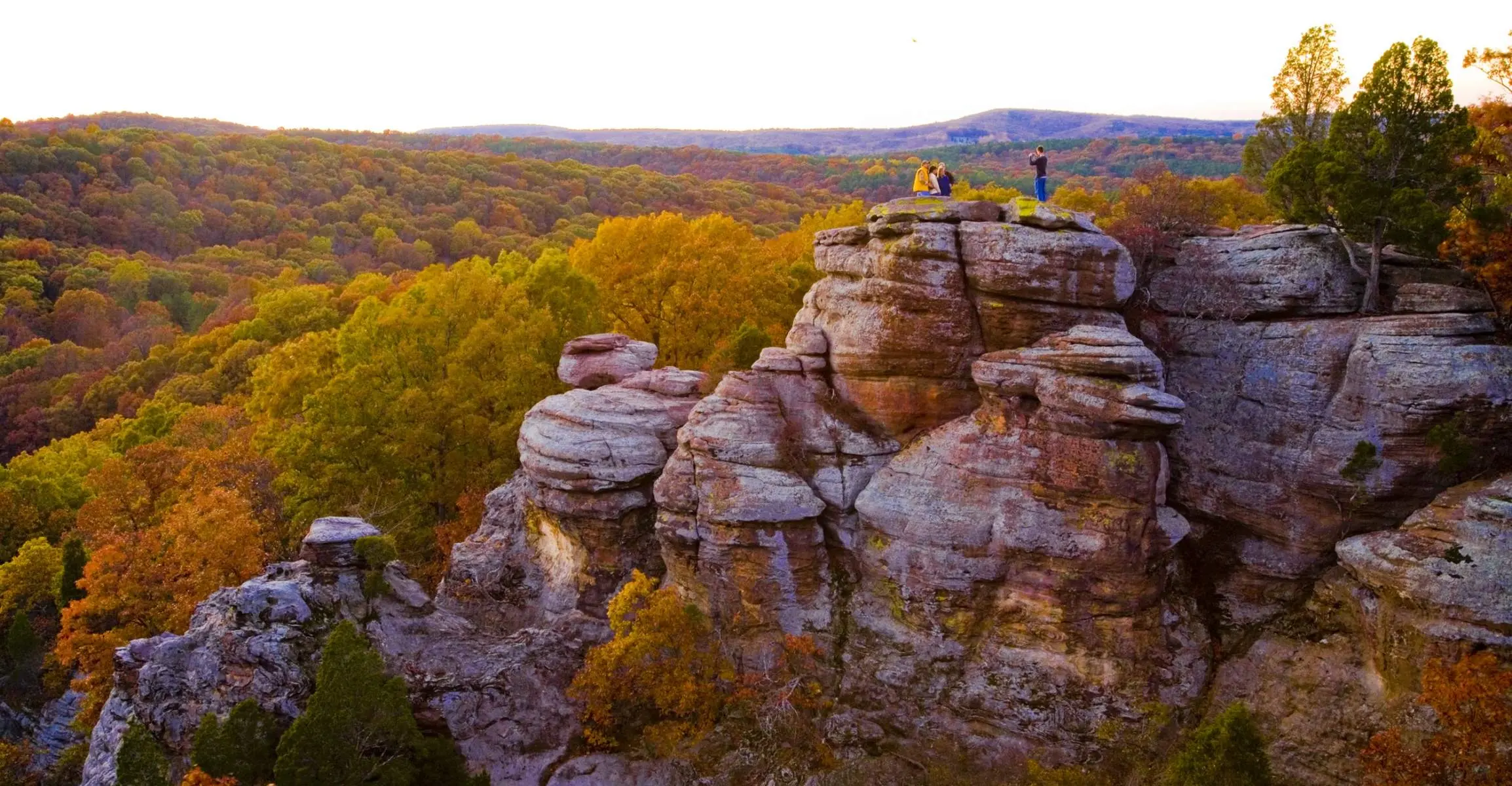 Un gruppo distante di quattro persone in posa per una foto in cima a una scogliera rocciosa, con vista su foreste di alberi verdi e dorati