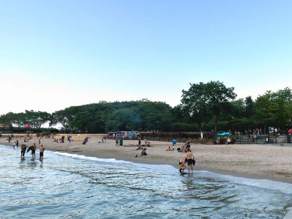 Piccoli gruppi di persone su una tranquilla spiaggia lacustre al tramonto, con bassi alberi verdi alle spalle