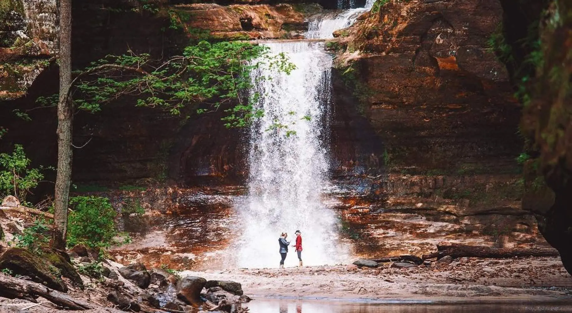Due persone in piedi davanti a una cascata, accanto a un laghetto sereno.