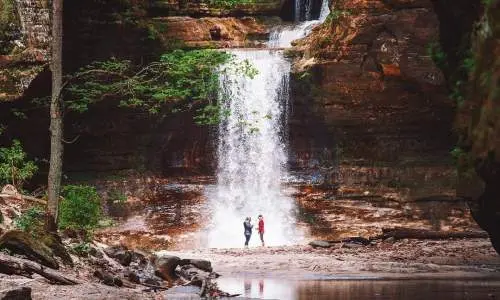 Due persone in piedi davanti a una cascata, accanto a un laghetto sereno.