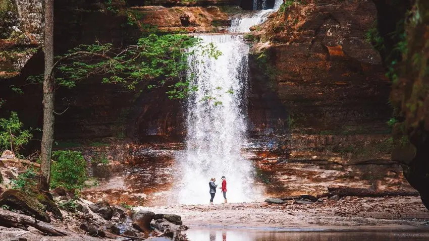 Due persone in piedi davanti a una cascata, accanto a un laghetto sereno.