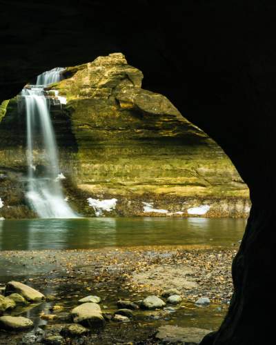 Guardando verso l'esterno di una grotta, una cascata si getta in una piscina, accanto a pareti di roccia innevate