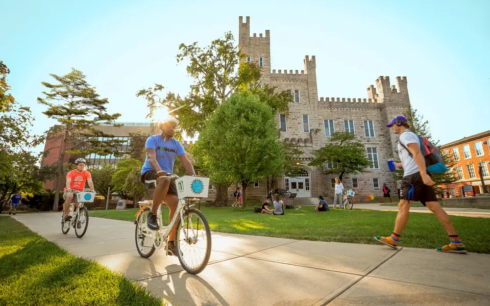Due persone attraversano in bicicletta un campus universitario in una giornata di sole