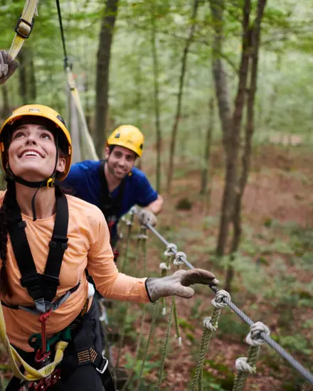 Due persone, munite di caschi e imbracature, si muovono lungo un percorso a corde sospese nel bosco.