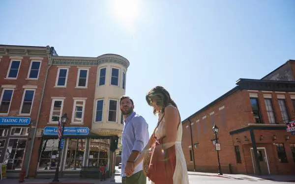 Un'inquadratura dal basso di una giovane coppia che cammina lungo la strada principale di una piccola città, con gli edifici in mattoni e il sole alle spalle
