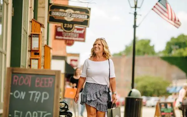 Una donna cammina lungo il marciapiede della strada principale di una piccola città, guardando la vetrina di un negozio. Le insegne di un negozio vecchio stile e una bandiera americana sono appese sopra