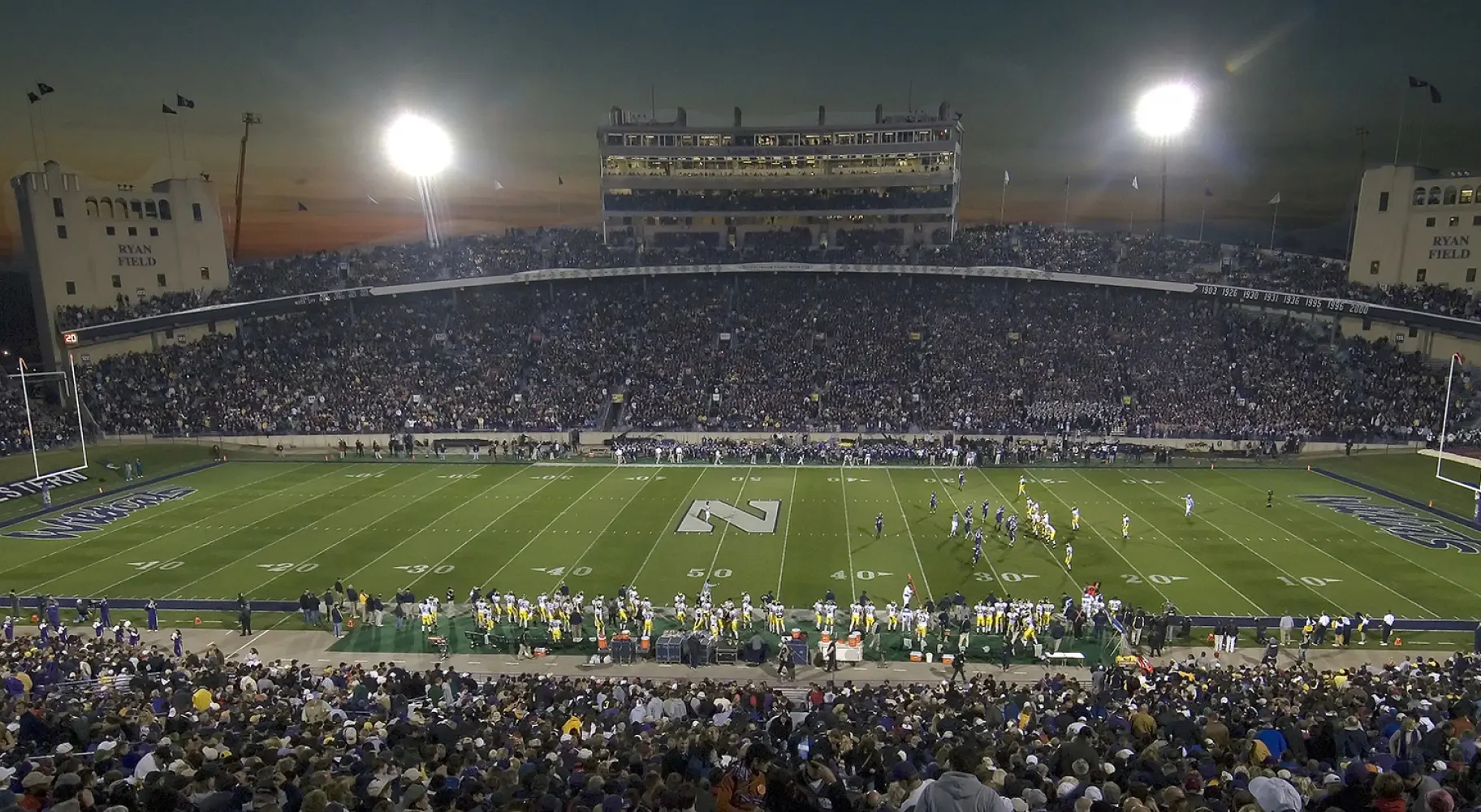 Un grande stadio di football universitario di notte prima di una partita, con gli spalti pieni di spettatori