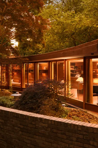 Moderna casa con vetrate curve illuminata al tramonto, circondata da alberi e da un muretto di mattoni.