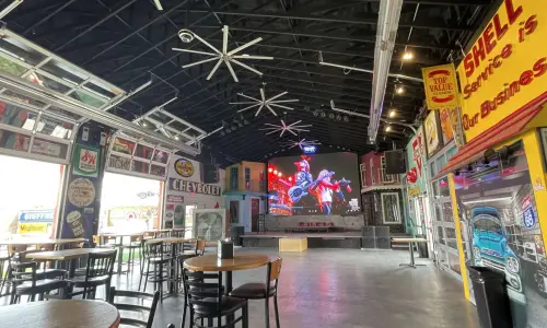 Interno di un bar spazioso e locale con musica dal vivo, dotato di tavoli, insegne e arredi vintage ispirati al mondo dell'automobilismo, oltre a un grande schermo sopra il palco.